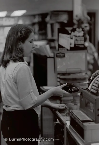 Black-and-white image of a woman handing a 35mm camera to a customer near a cash register