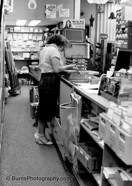 Woman in white blouse and dark skirt writing at a cluttered camera shop counter lined with boxes