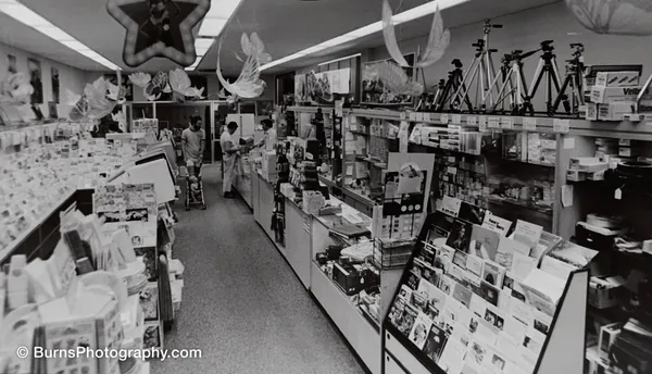 Camera store interior with glass counters, tripods on a high shelf, and paper decorations hanging from the ceiling