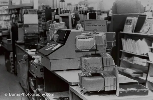 Black and white store interior: new cash register on a counter, wire racks of photo albums, sign reading Snapshot Books