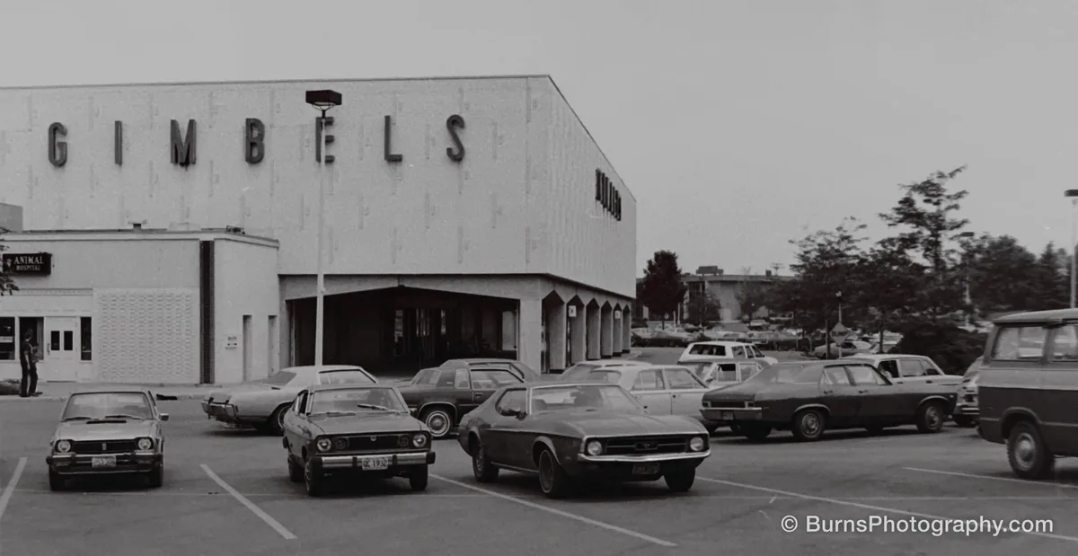 Black and white view of a department store with large GIMBELS letters above a parking lot of 1970s cars