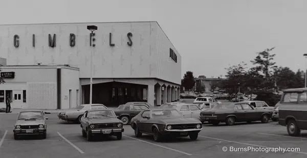 Black and white view of a department store with large GIMBELS letters above a parking lot of 1970s cars