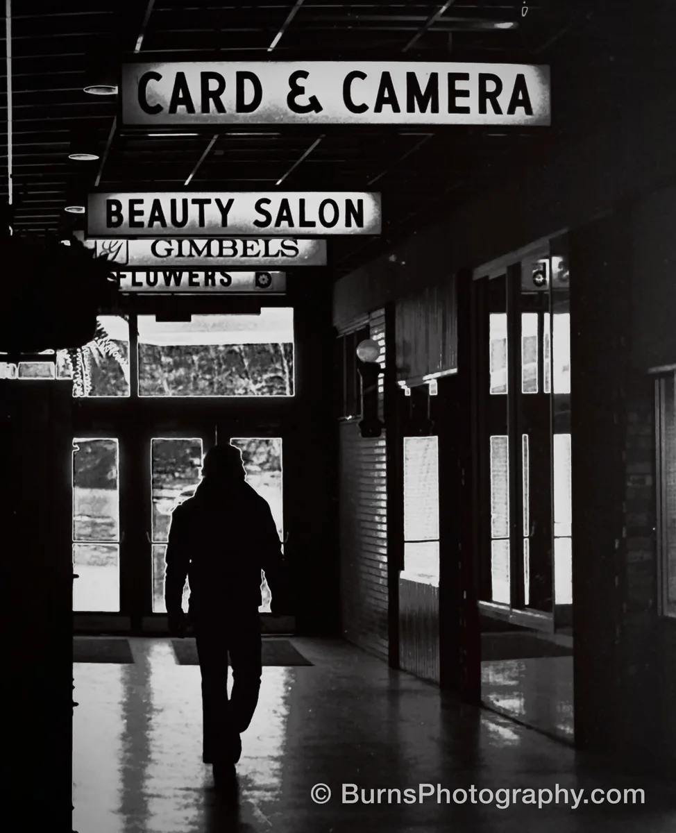 Black and white interior of a shopping mall with signs and a silhouetted person walking down the mall