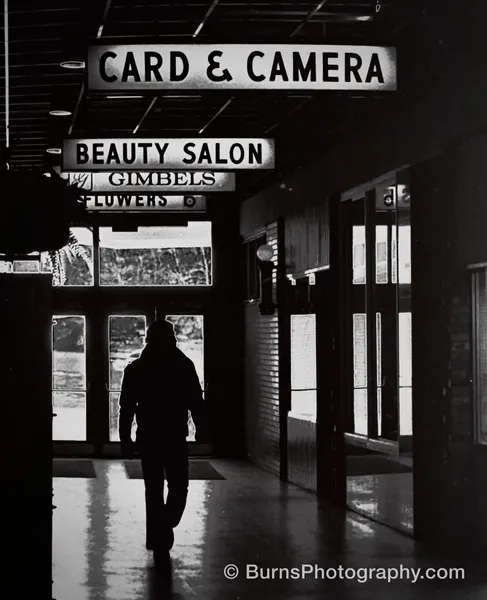 Black and white interior of a shopping mall with signs and a silhouetted person walking down the mall