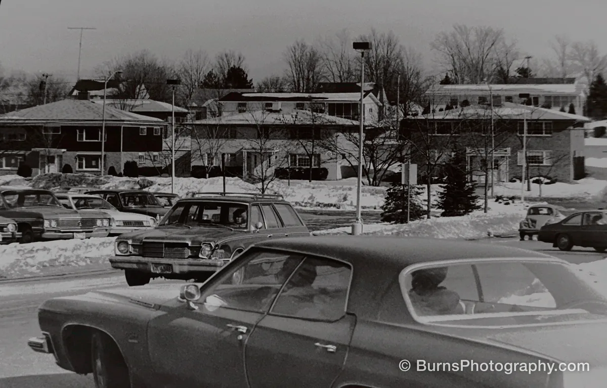 Black and white scene of 1970s cars in a snowy Hilldale parking lot with brick apartment buildings behind