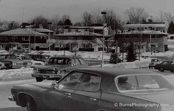 Black and white scene of 1970s cars in a snowy Hilldale parking lot with brick apartment buildings behind