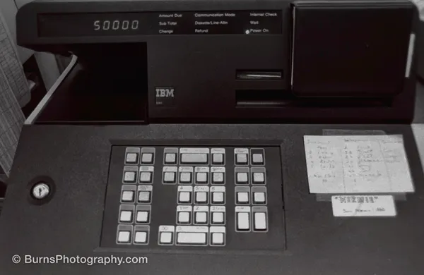 Black IBM cash register terminal with numeric keypad, lit display reading 50000, and taped handwritten note