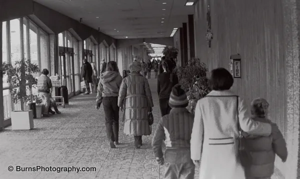 Black and white view down an enclosed Hilldale shopping mall corridor with people in winter coats walking to stores.