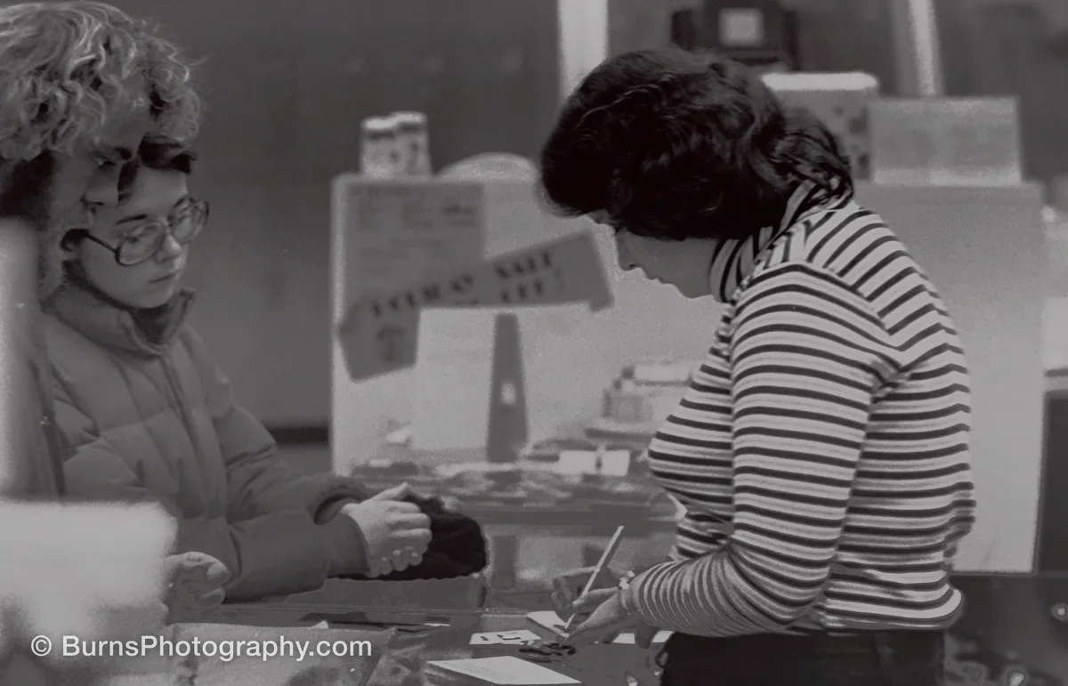 Black and white indoor scene: woman filling out a print order form at a counter, customers watching