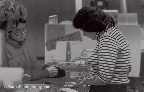 Black and white indoor scene: woman filling out a print order form at a counter, customers watching