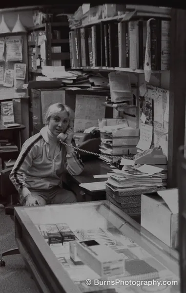 Woman in track jacket on a rotary phone at a cluttered camera store counter with shelves of boxes