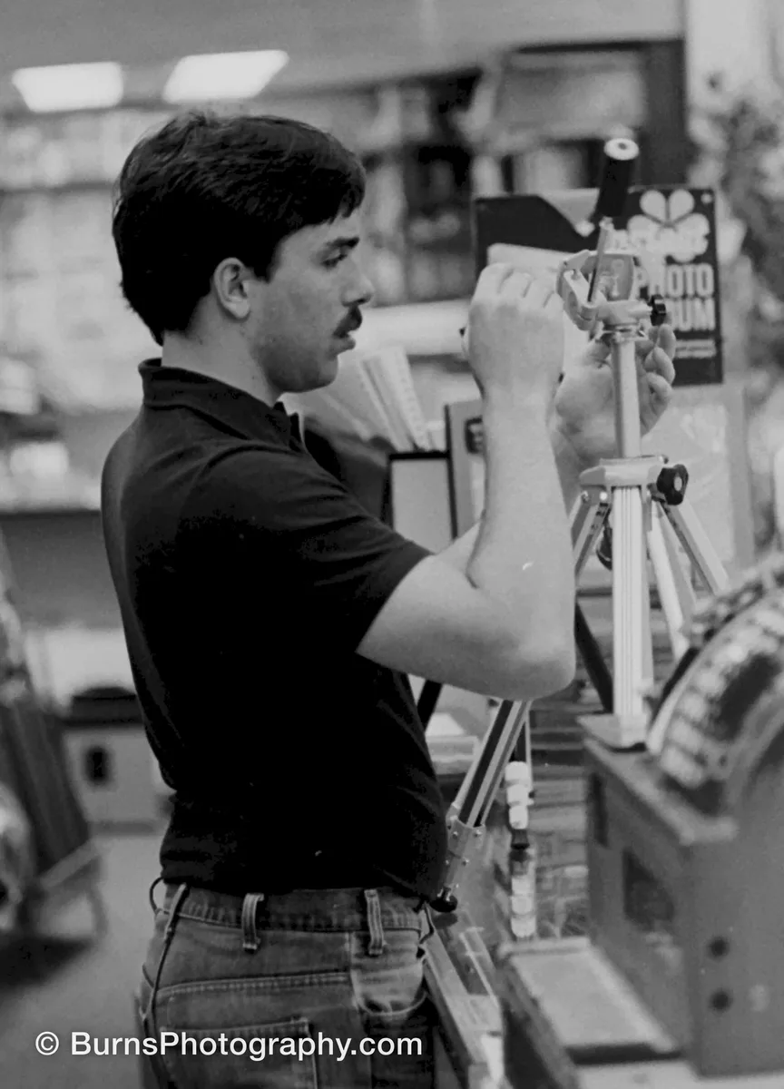 Black-and-white image of a man in a dark polo adjusting a tripod head in a camera store