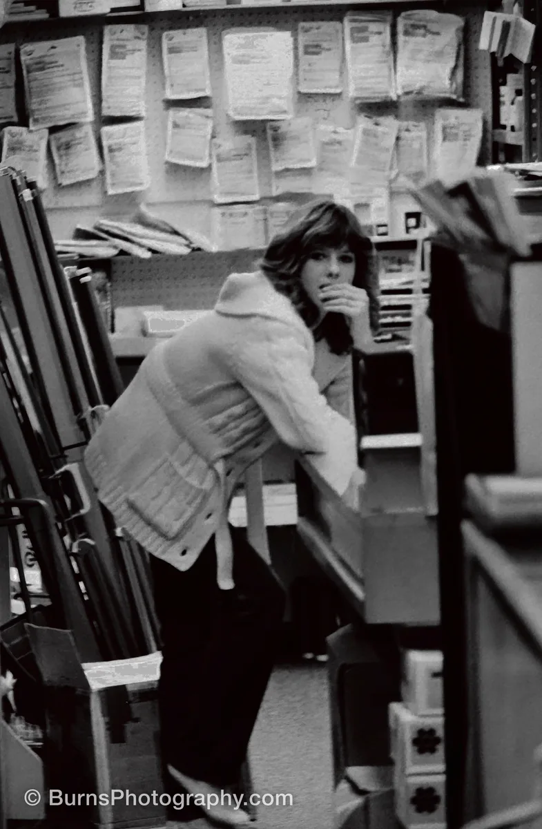 Black and white photo of a woman in a cable-knit cardigan leaning over a shop counter beneath a wall of darkroom chemicals and photo papers.