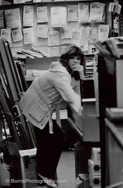 Black and white photo of a woman in a cable-knit cardigan leaning over a shop counter beneath a wall of darkroom chemicals and photo papers.