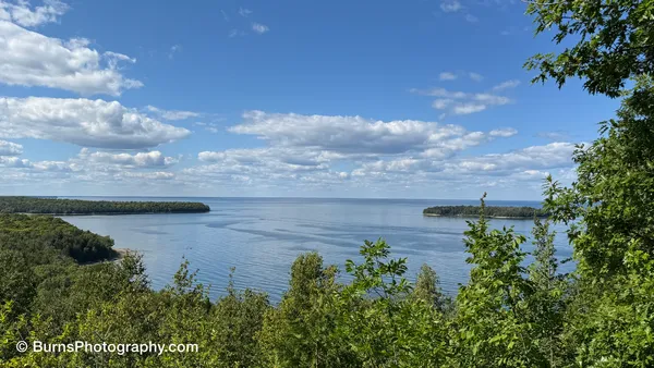Elevated view over a blue bay framed by green treetops, wooded points and an island under scattered clouds