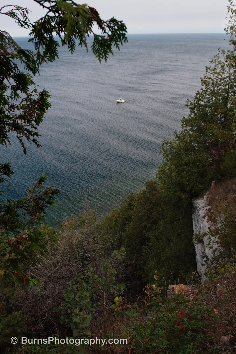 View from a wooded limestone bluff over calm water with a small white boat anchored offshore