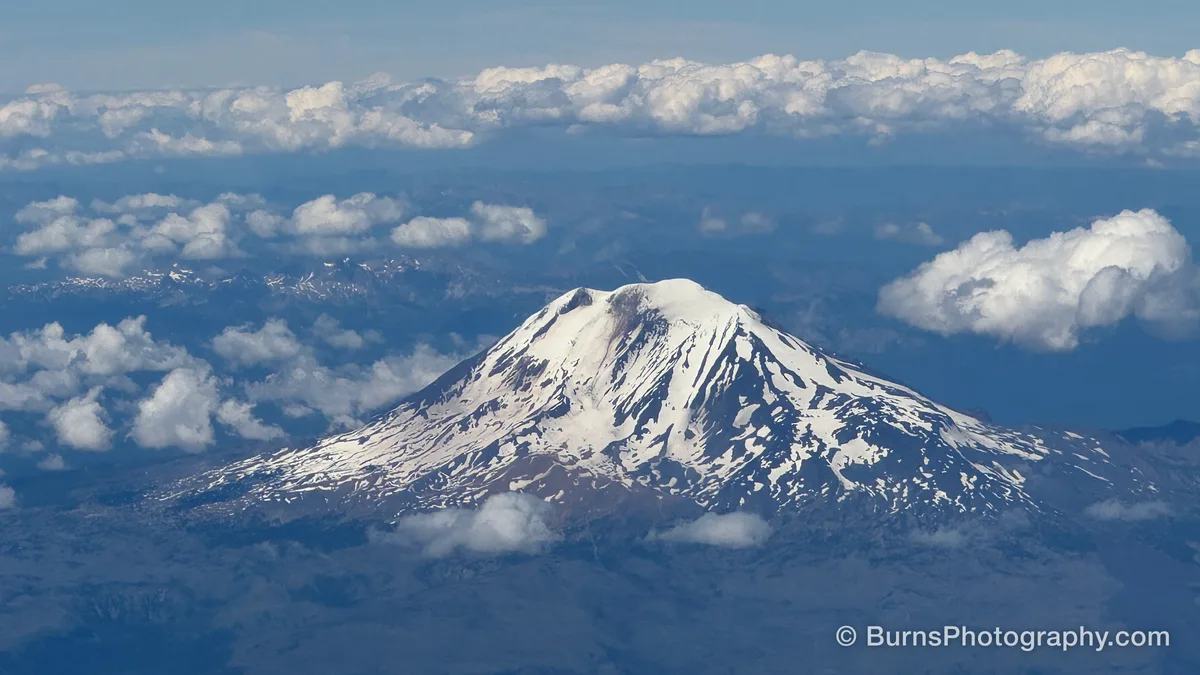Aerial view of a snow-streaked volcano rising above scattered white clouds with blue ridges behind