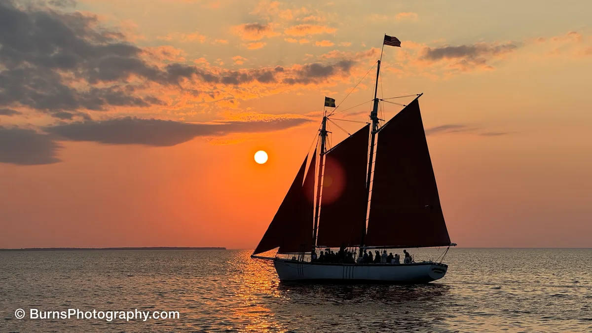 Silhouetted two-masted schooner with dark sails on calm water, sun low behind it in an orange sky