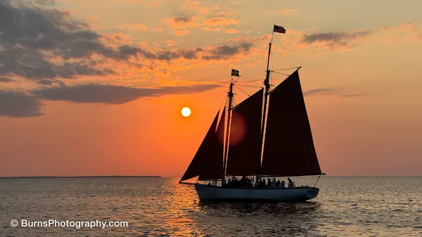 Silhouetted two-masted schooner with dark sails on calm water, sun low behind it in an orange sky