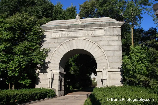 Granite memorial arch inscribed CAMP RANDALL with soldier statues, surrounded by green trees and hedges