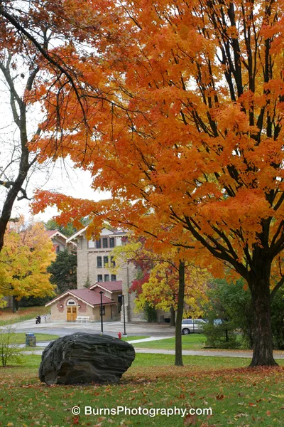 Orange maple tree over a lawn with a glacial boulder, limestone building with red tile roofs behind