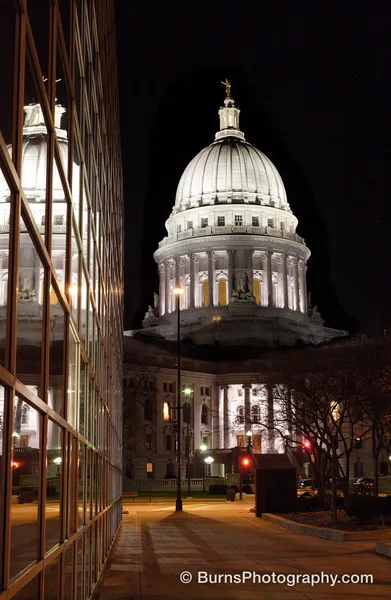Floodlit Wisconsin State Capitol dome at night, reflected in a glass office facade beside an empty sidewalk