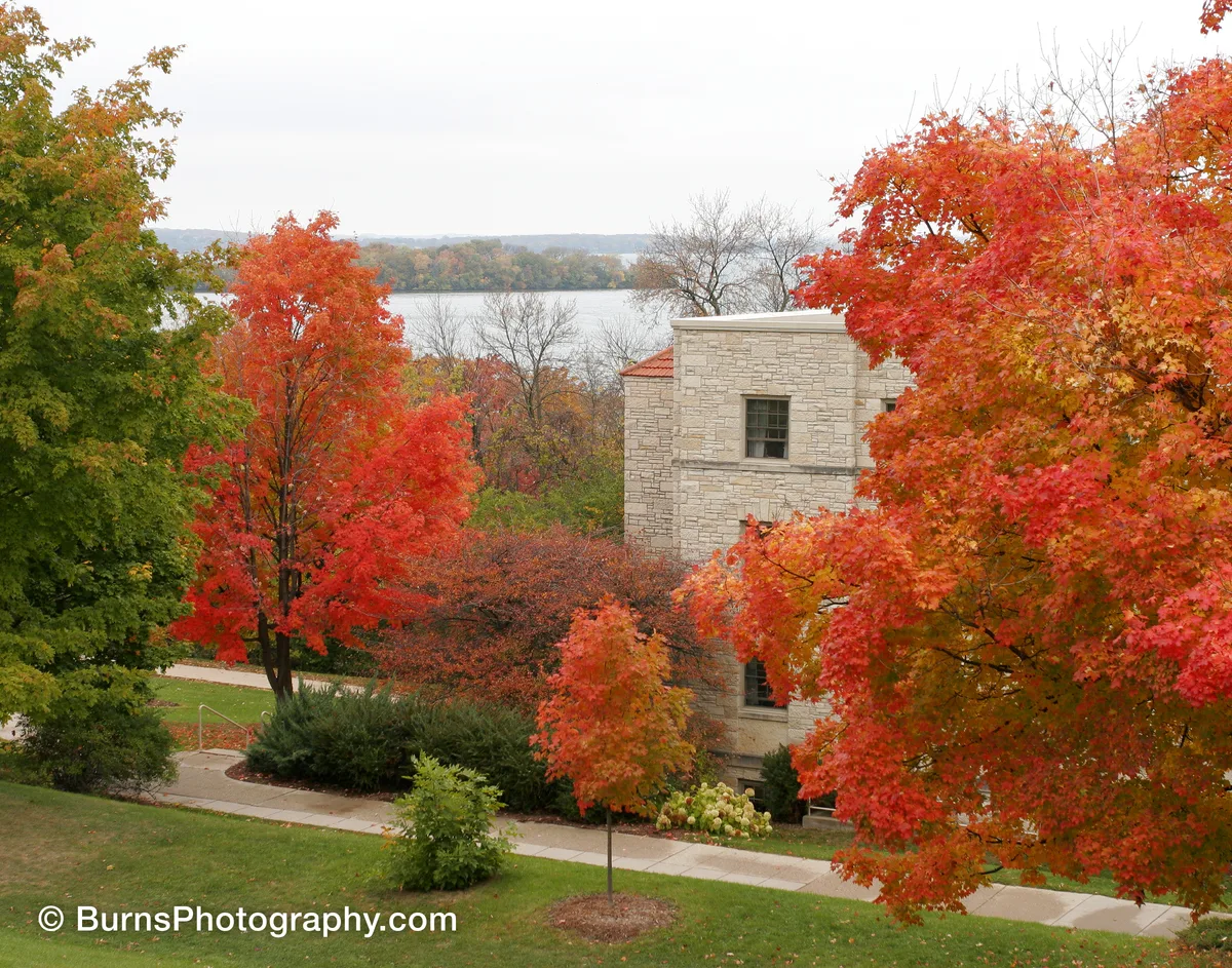 Red and orange maples around a limestone building on a slope, with a gray lake and wooded shore behind