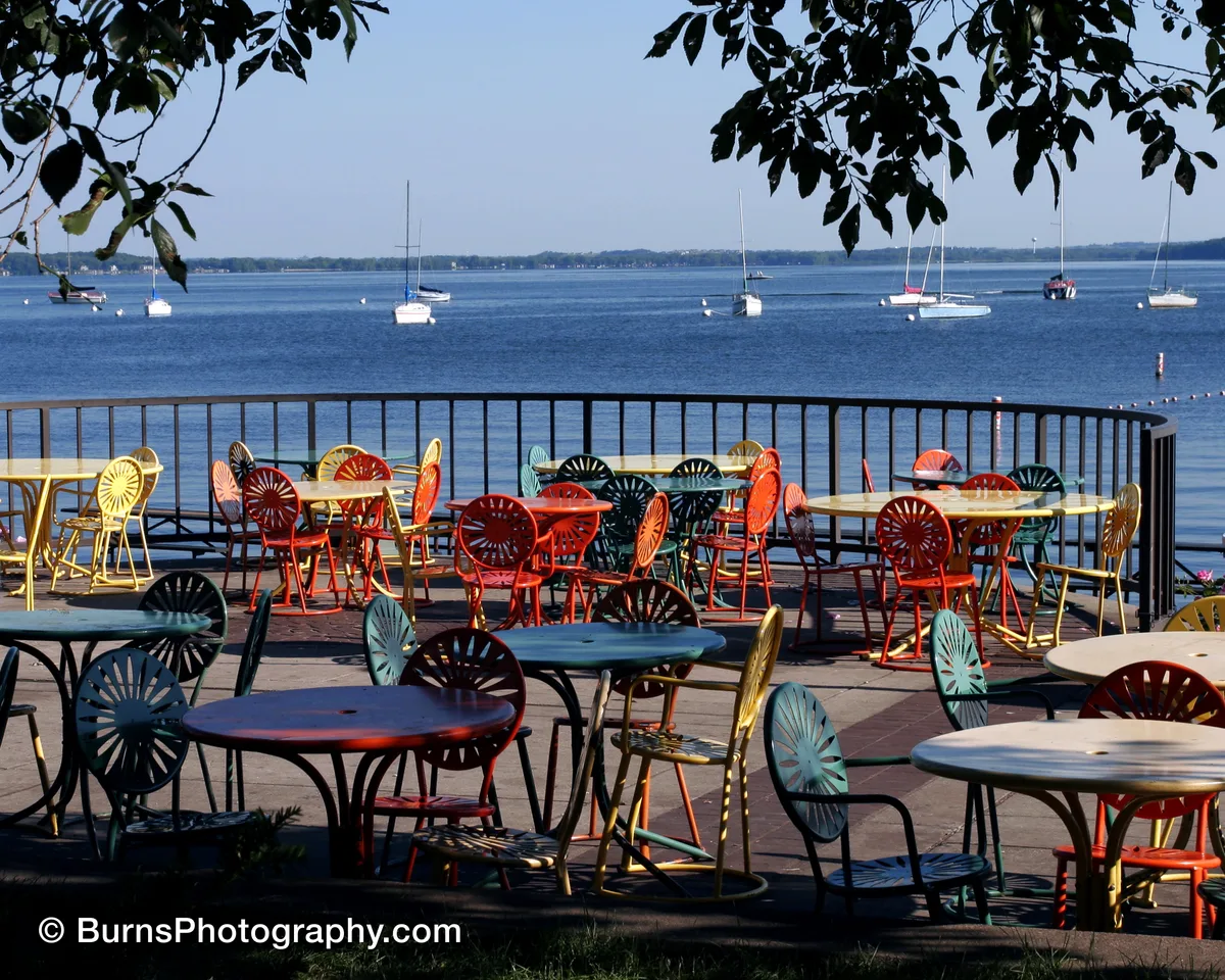 Colorful sunburst metal chairs and tables on a lakeside terrace with moored sailboats beyond a railing