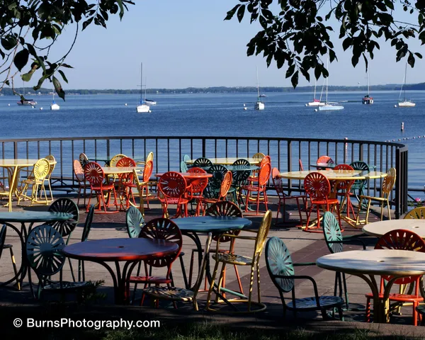 Colorful sunburst metal chairs and tables on a lakeside terrace with moored sailboats beyond a railing
