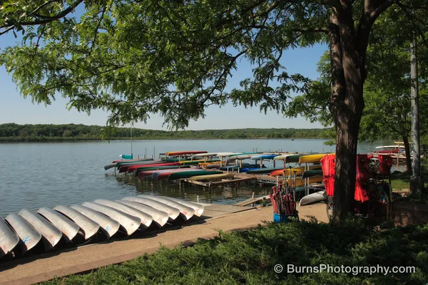 Overturned aluminum canoes on a beach beside docks of colored canoes and red life jackets under a leafy tree
