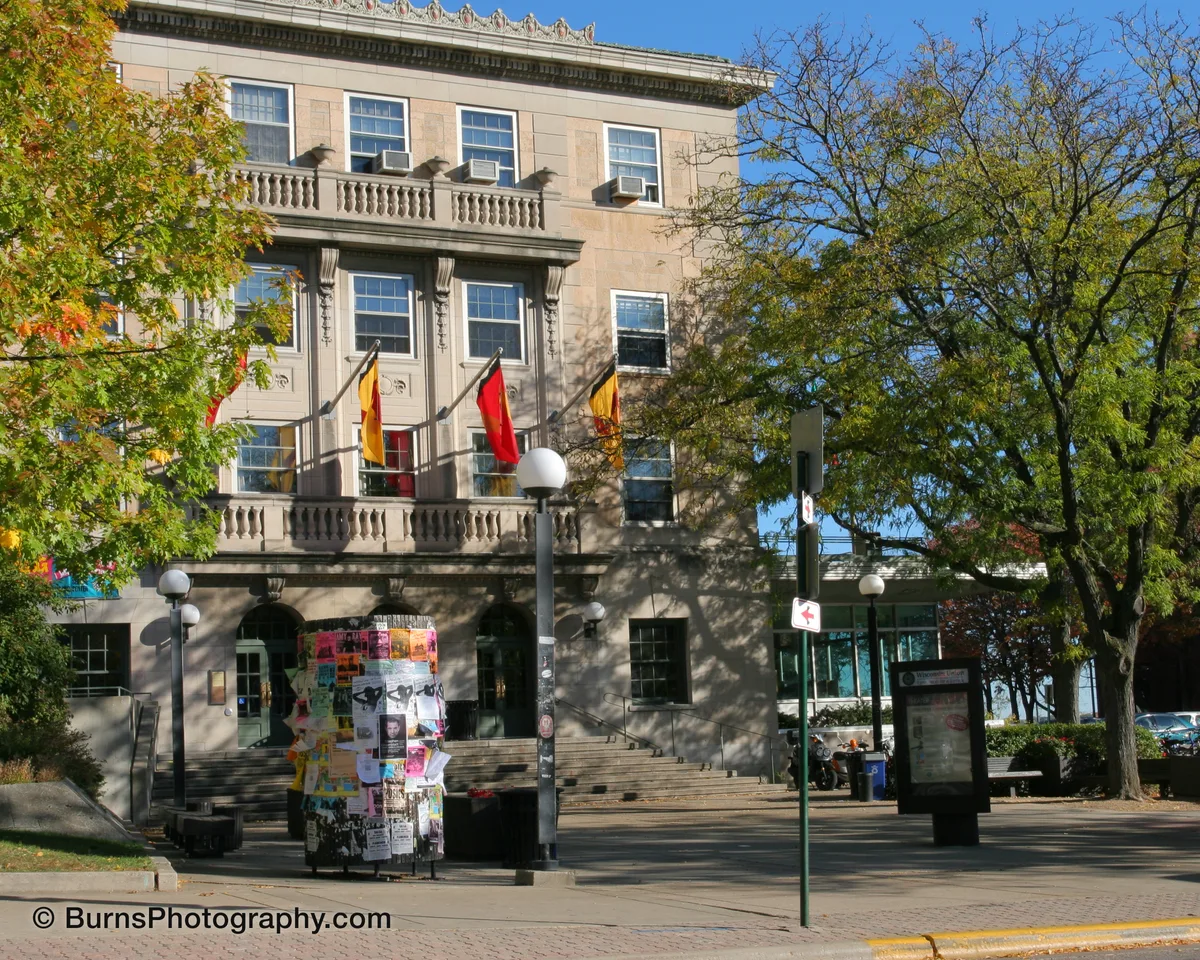 Limestone university union building with red and yellow banners, poster-covered kiosk, and autumn trees