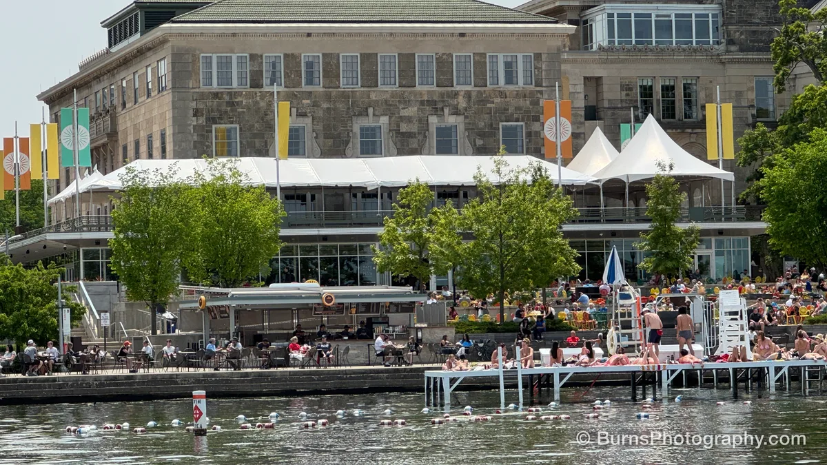 Crowded lakeside terrace with white tents, colorful chairs, swim pier and swimmers in Lake Mendota