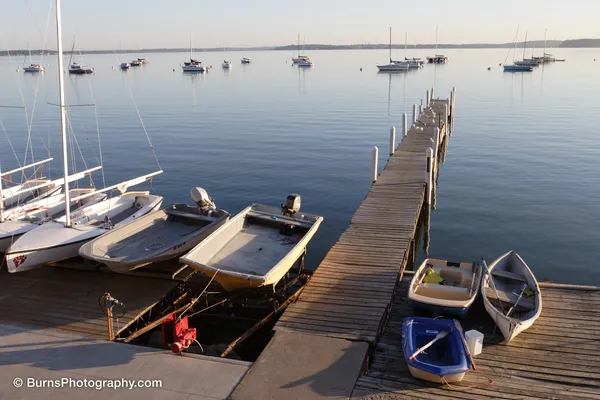 Wooden pier extending into a calm lake with moored sailboats, dinghies, and small boats in morning light