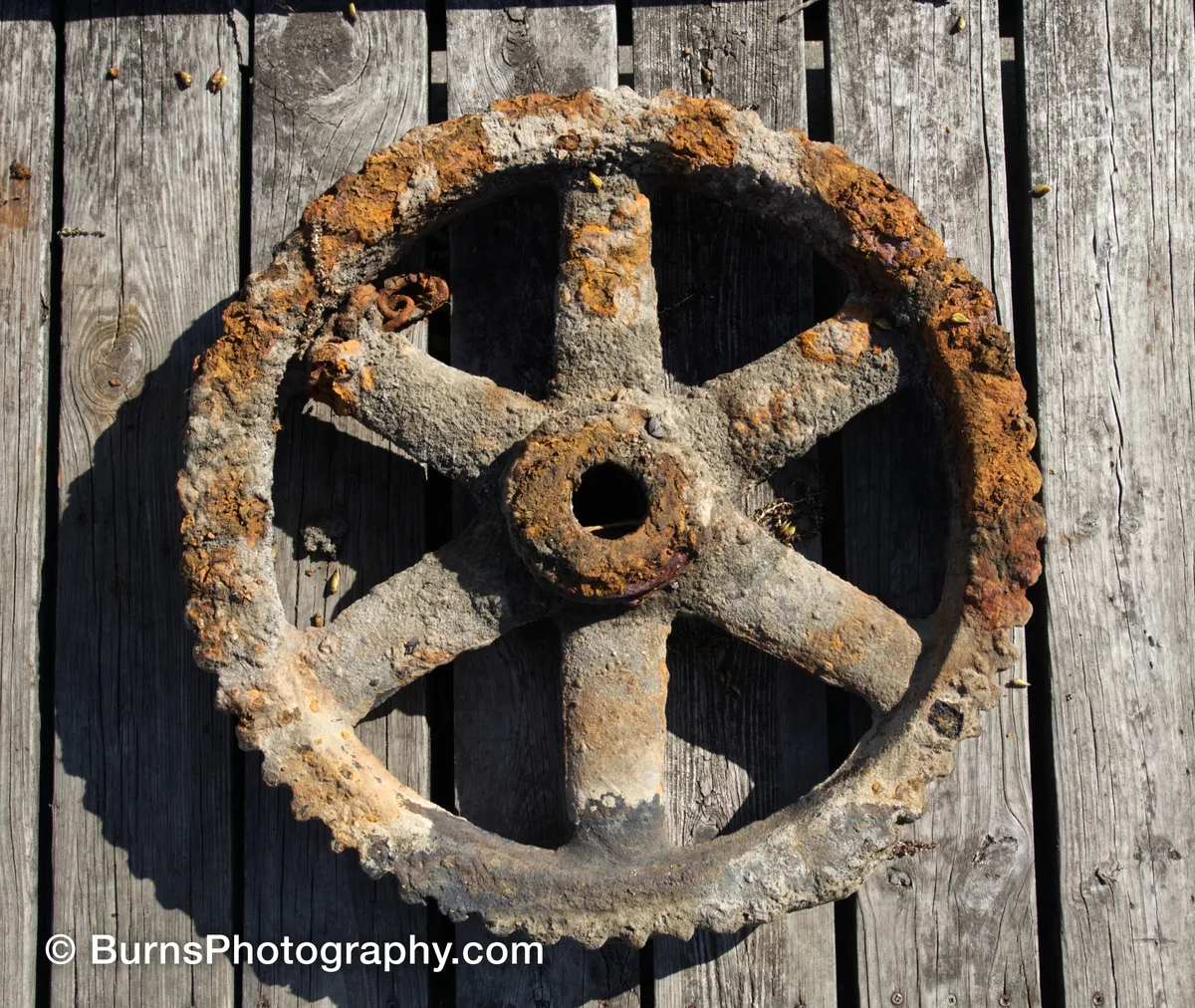 Rusted six-spoke iron gear with toothed rim lying on weathered gray wooden planks in sunlight
