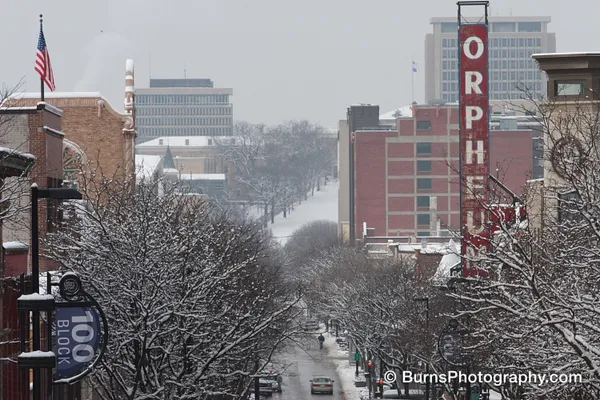 Snowy downtown street lined with bare trees, vertical Orpheum theater sign and brick buildings under gray sky