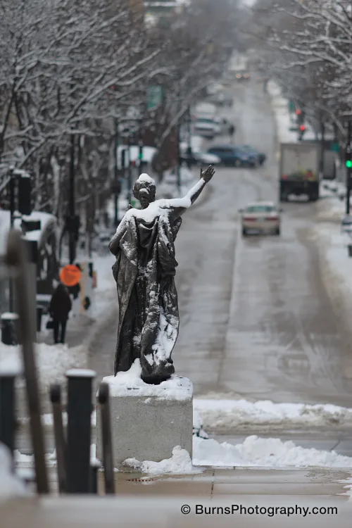 Bronze robed statue with raised arm, covered in snow, on a pedestal above a slushy city street