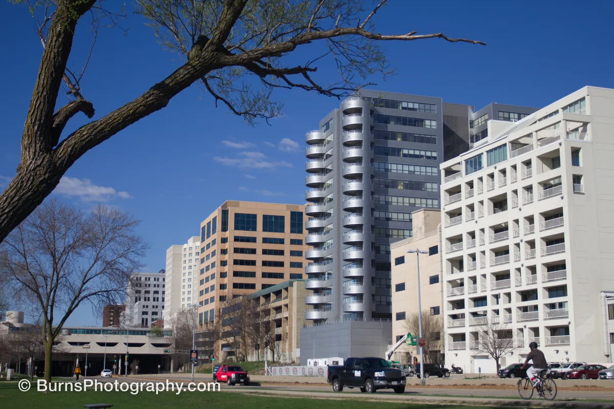 Downtown buildings with curved balconies under blue sky, bare tree branch at left, cyclist and trucks on street