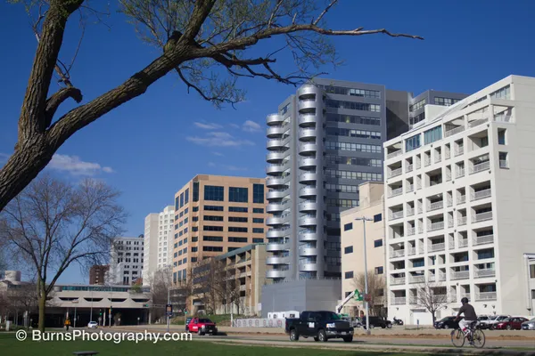 Downtown buildings with curved balconies under blue sky, bare tree branch at left, cyclist and trucks on street