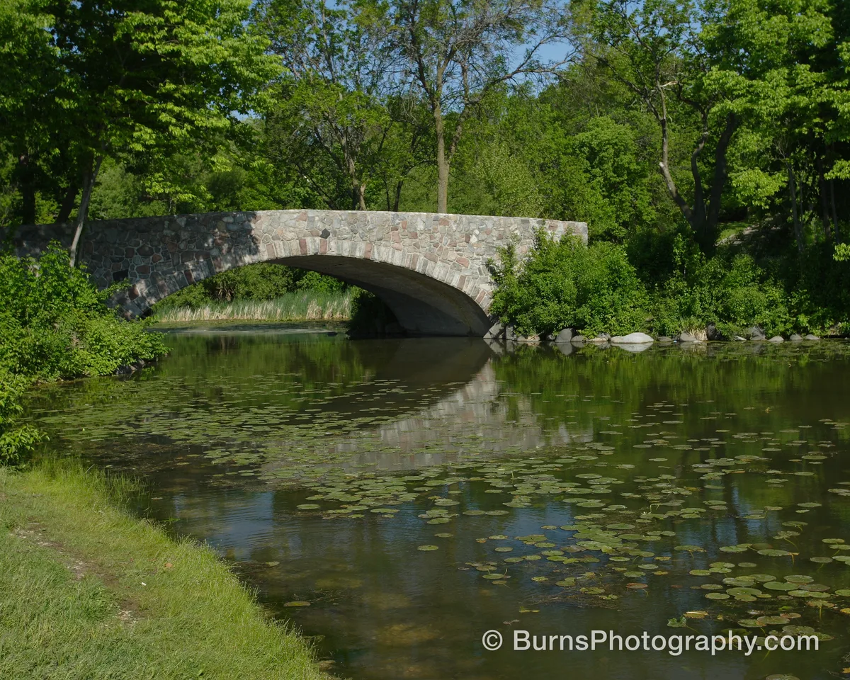 Stone arch bridge over a calm green lagoon covered in lily pads, surrounded by leafy summer trees