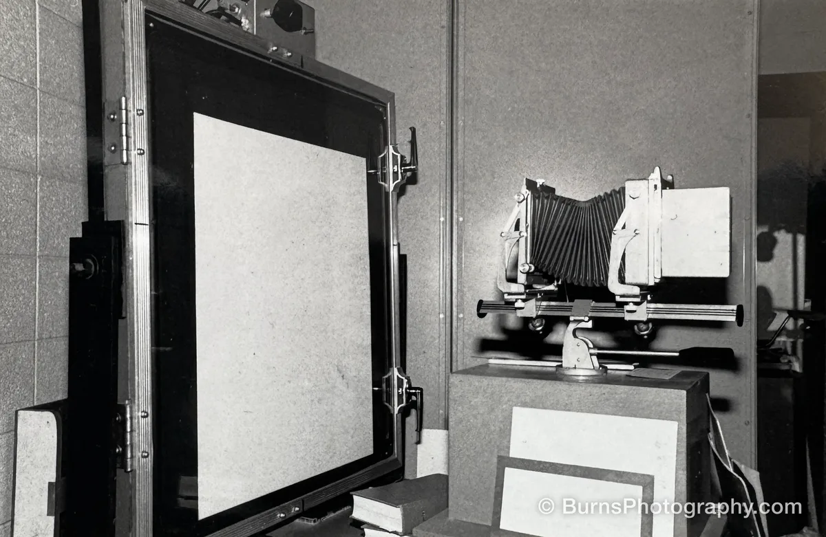 Black-and-white view of a bellows view camera on a rail stand facing a large hinged copy easel in a lab