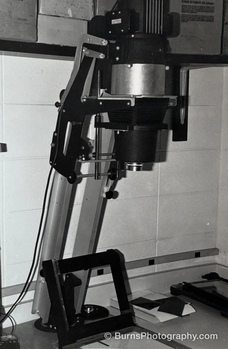 Black-and-white photograph of a large photographic enlarger on a tilted column beside a concrete block wall