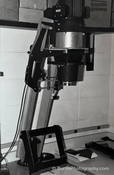 Black-and-white photograph of a large photographic enlarger on a tilted column beside a concrete block wall