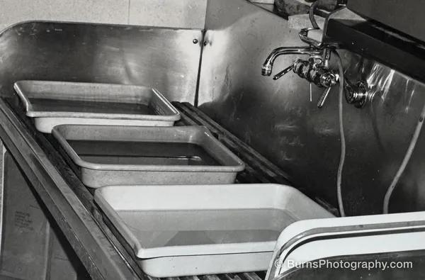 Three photographic developing trays lined up inside a stainless steel sink with wall-mounted faucets