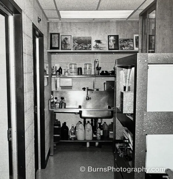 Chemical mixing interior with stainless sink, shelves of bottles and jugs, framed prints above