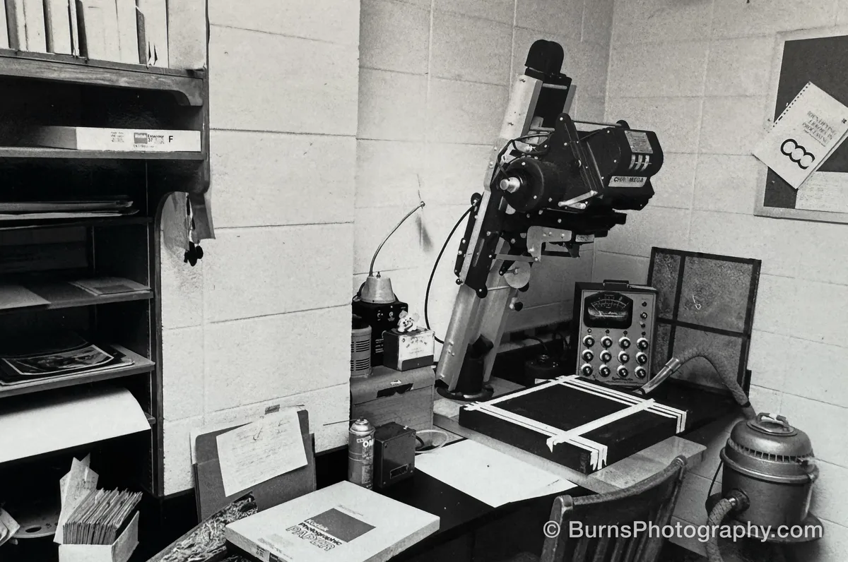 Color darkroom scene with a tilted enlarger, timers, easel, paper boxes and shelving