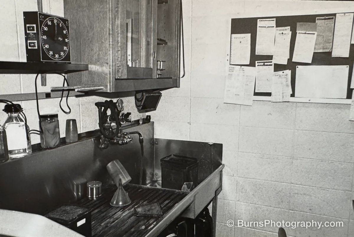 View of a film darkroom sink with funnel, graduates, timer on a shelf, and a corkboard of printed instructions