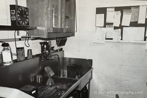 View of a film darkroom sink with funnel, graduates, timer on a shelf, and a corkboard of printed instructions